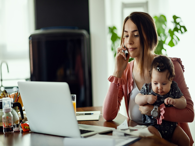 Woman sitting at her desk, on the phone, holding a child, looking at a laptop